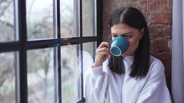 Beautiful Happy Caucasian Brunette Sitting Next to the Window Drinking Tea and Enjoying Her Day