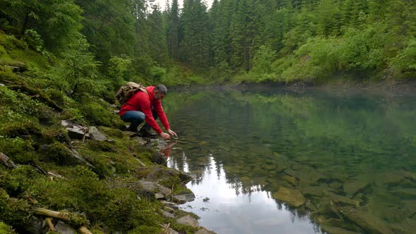 A Bearded Man with a Backpack Drinks Water From a Clean Lake