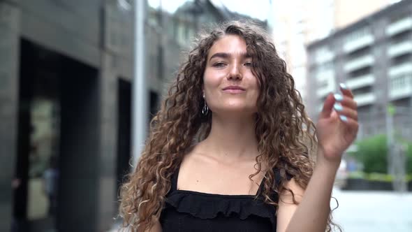 a Young Woman with Curly Hair Poses on a City Street, Raising and Lowering Her Hands. the Camera