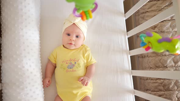 Beautiful Little Girl in a Yellow Bodysuit Lies in a Crib and Examines Toys