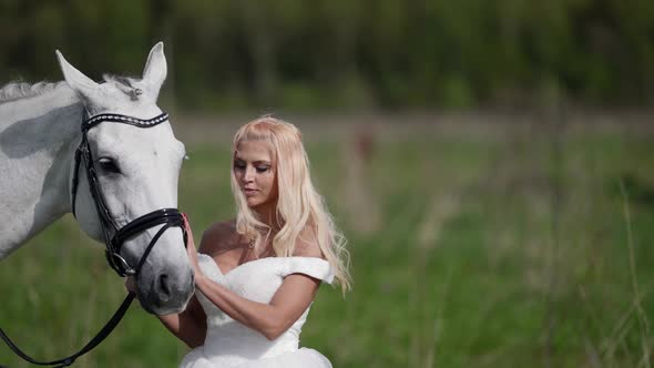 Romantic Blonde in White Wedding Dress is Communicating with Horse at Nature Portrait Shot