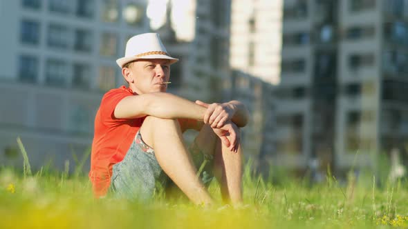 Man in hat is resting while sitting on the grass in a city park, tracking the camera