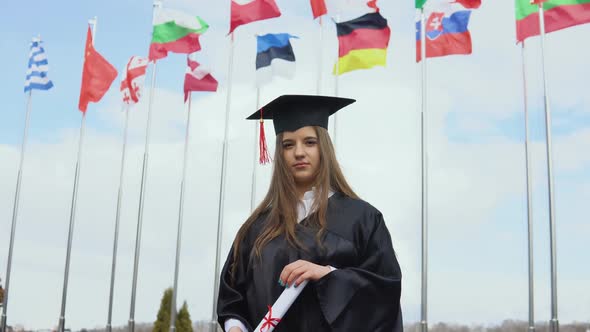 A University Graduate Stands on the Background of Flags of the World Holding Graduatind Diploma of