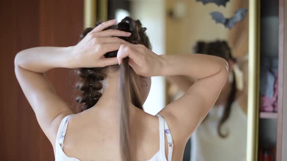 young woman weaves her braid in front of a mirror