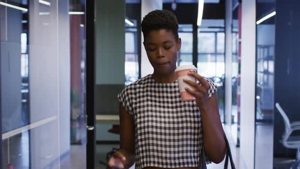 African american businesswoman using smartphone holding coffee walking in office corridor