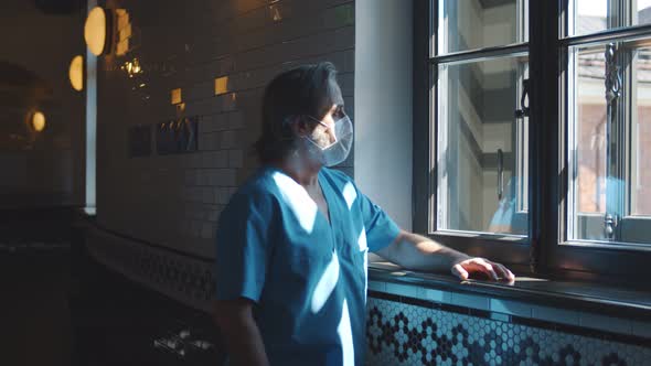 Senior Doctor in Uniform and Medical Mask Resting Looking Through Window in Hospital Corridor