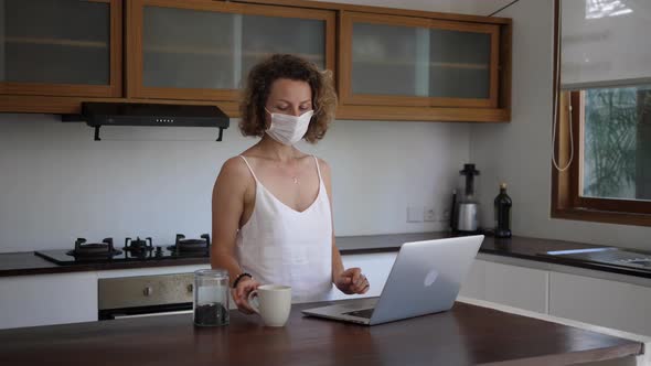 Young Woman in Protective Mask Works on a Laptop and Drinking Tea at Her Kitchen