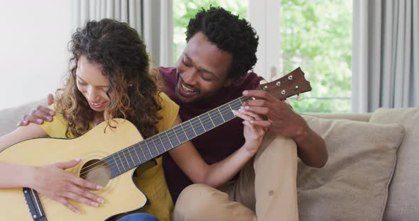 Happy biracial couple sitting on sofa together and playing guitar