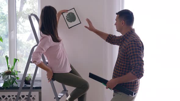 Home Renovation, Beautiful Lovers Hang Picture and Shelf on White Wall Using Ladder in Apartment