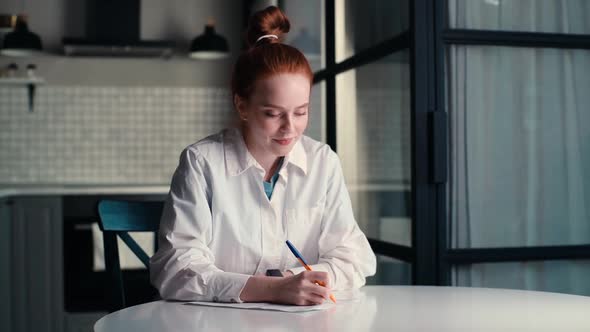 Portrait of Dreamy Young Woman Writing Notes in Paper Notebook Using Pen Sitting at Desk at Home