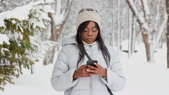 Young Black Woman in White Jacket Uses Her Phone in Winter Forest