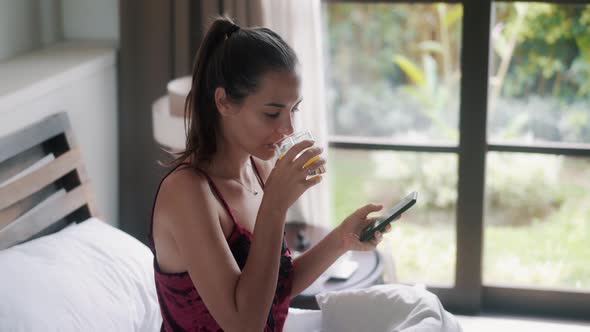 Girl with Juice Surfs Internet with Smartphone on Large Bed