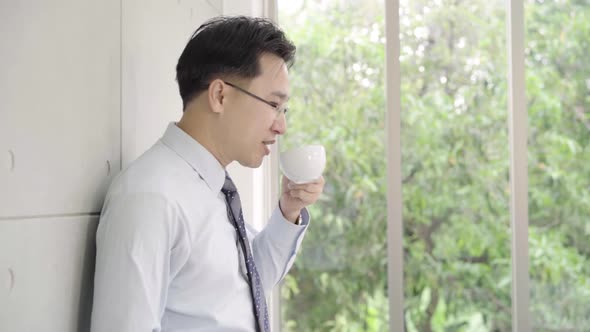 Mature asian businessman drinking a coffee and looking out of a window at the city