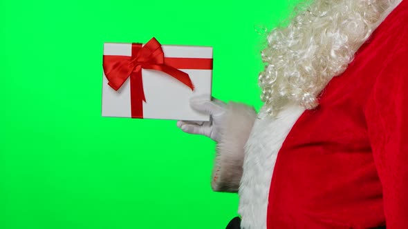 Side View of Santa Claus with Beard in White Gloves and a Red Suit Twirls a Gift Box in His Hands