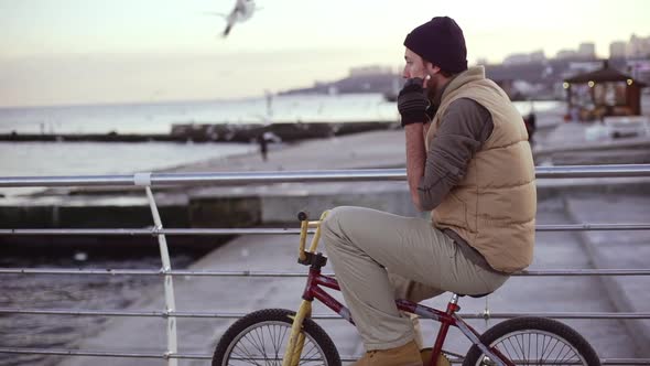 Young Caucasian Male in Sock Cap Sitting on Bmx Bicycle Watching Sea and Gulls Then Putting on