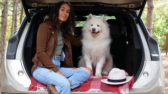 A young woman mixed-race in a stylish hat sits with a dog in the trunk, in the woods.