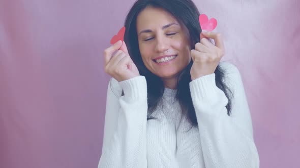 Smiling Funny Young Brunette in a White Sweater Smiles Blinks and Waving Red Hearts on a Pink