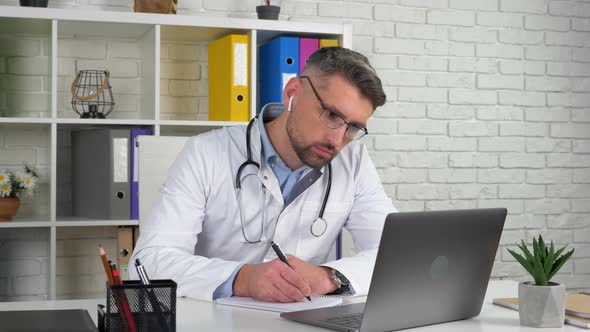 Doctor wears white medical coat in hospital office using laptop writes notebook