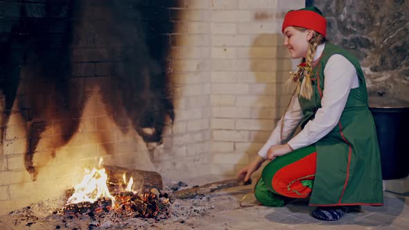 Pretty female elf is sitting near fire and rubbing her hands indoors.