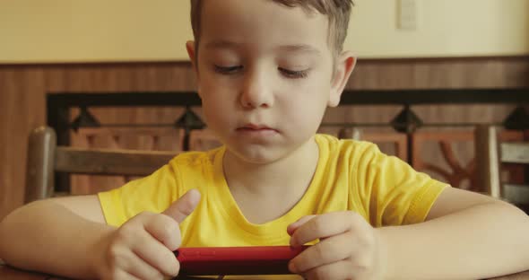 Portrait of a Little Boy Cute Child with a Looking at the Phone While Sitting at the Table a