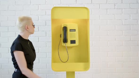 Blonde Woman with Short Haircut Picks Up Phone in Yellow Telephone Booth and Talks on Phone