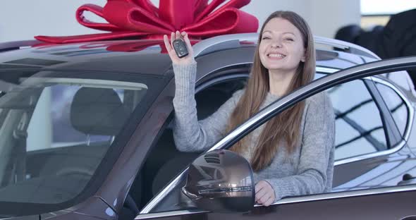 Young Caucasian Woman Standing at New Automobile in Car Dealership with Keys and Smiling. Portrait