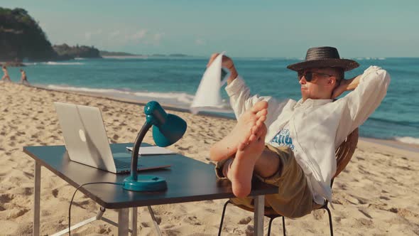 Young Caucasian Man Freelancer Sits on Beach at Desk with Laptop and Lamp