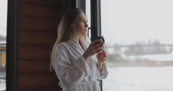 Young Beautiful Woman in White Bathrobe Drink Coffee By the Window at Home in the Morning