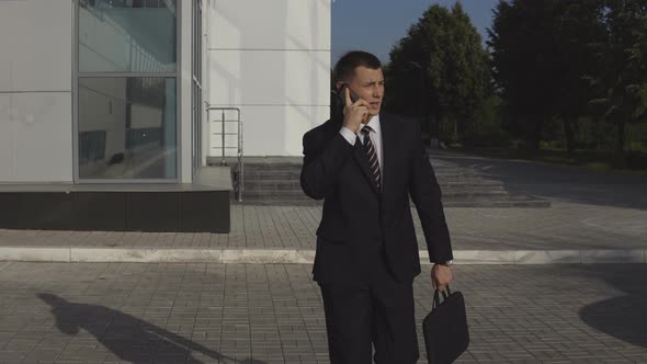 Serious Handsome Businessman in Black Suit with Diplomat Talking on Smartphone and Hurrying To