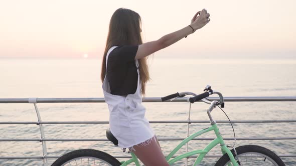 Young Attractive Woman Doing Selfie on a Bicycle at Sunrise or Sunset Near the Sea