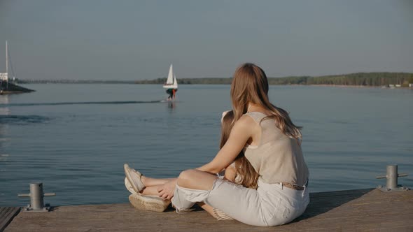 Woman with Daughter Sit on a Pier Look at Man Surfing on a Lake at the Sunset