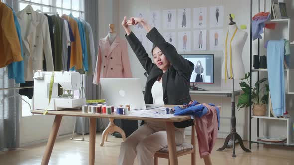 Asian Female Designer In Business Suit Working On A Laptop And Stretching While Designing Clothes 