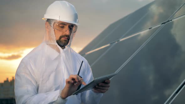 Thoughtful Solar Worker Is Standing with a Computer Beside a Solar Installation in the Sunset