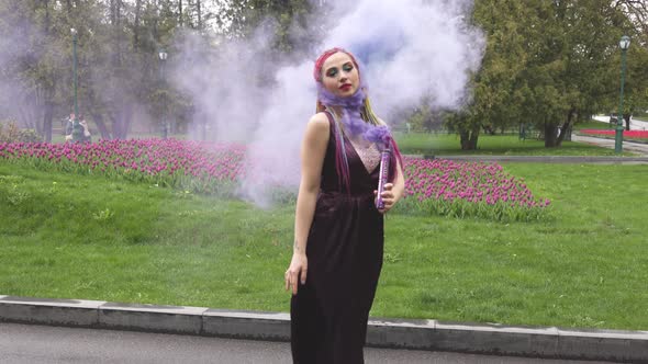 A Girl with a Smile with Bright Makeup and Colored Braids in a Purple Dress Blows Artificial Purple