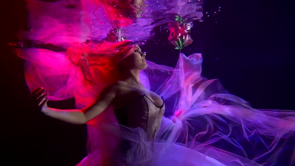a Woman with Red Hair in a Corset Floats in the Dark Under Water with a Translucent Fabric. Side