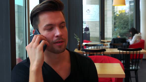 A Young Handsome Man Sits at a Table in a Cafe and Talks on a Smartphone - Closeup on the Face