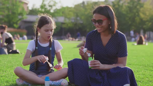 Caucasian Preteen Daughter and African Mother Blow Soap Bubbles on Blurry Background in Park