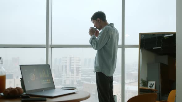 Pensive Young Man Drinking Coffee Looking at City From Apartment Window