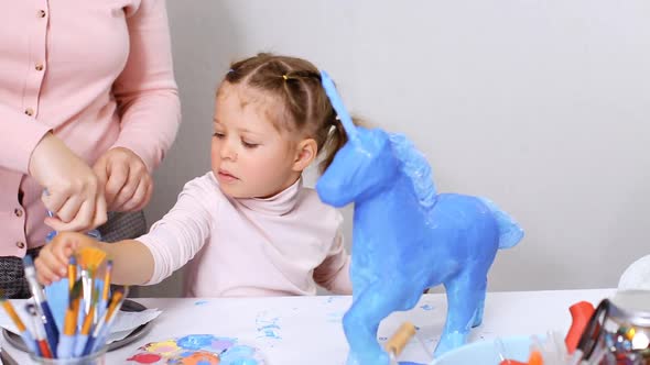 Step by step. Mother and daughter painting paper mache unicorn with blue paint together.