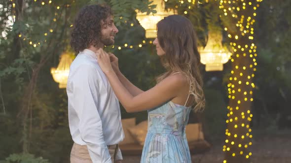 Loving Young Woman Adjusting Shirt of Handsome Caucasian Curly-haired Man and Smiling. Side View