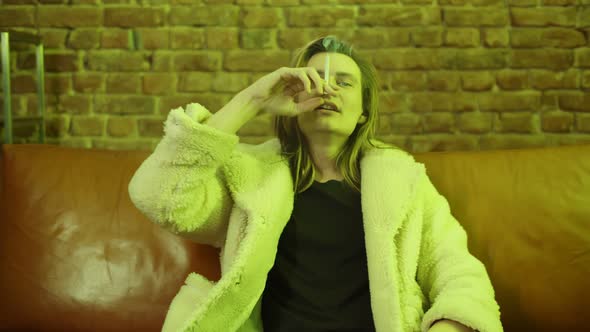 Portrait of Young Man Drug Dealer Sitting on Couch in Coat and Smoking