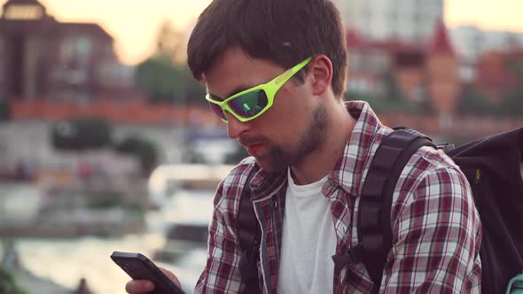 Young Caucasian Man Sitting on Fence on River Embankment Uses Smartphone Chatting While Waiting for