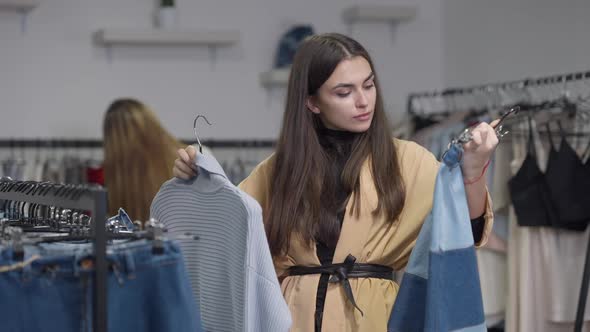 Gorgeous Young Woman Choosing Clothes in Shop Looking at Camera with Confused Facial Expression