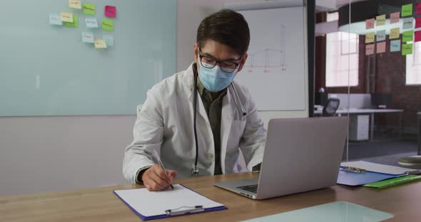 Asian male doctor wearing mask sitting at desk in meeting room making notes