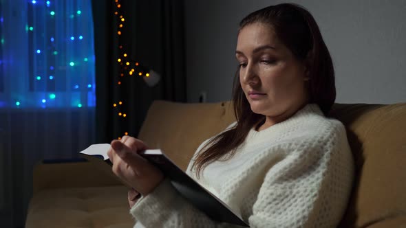 Young Woman Reads Book Sitting on Sofa in Room in Evening