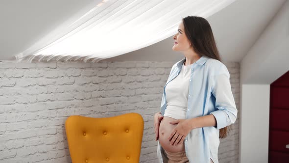 Smiling Beautiful Pregnant Young Woman Stroking Big Naked Belly Looking Out Window at Loft Interior