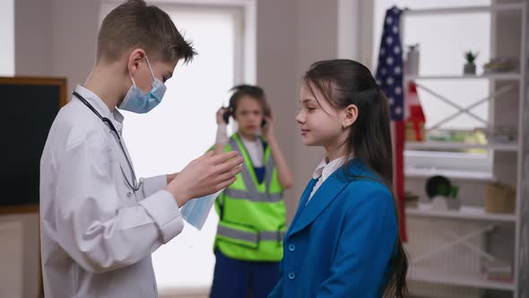 Side View Serious Teenage Boy in Medical Gown Putting Coronavirus Face Mask on Girl