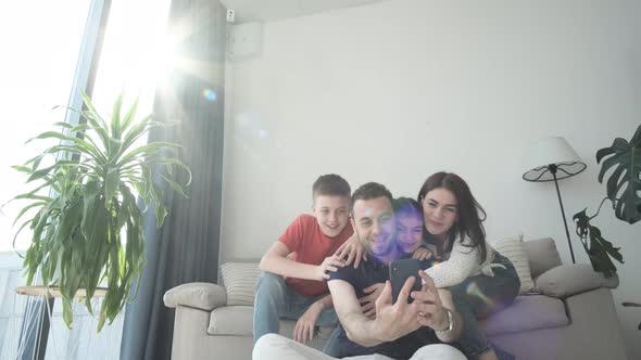 Happy Parents and Young Children Laughing Using Gadget for Selfie Photo Relaxing Together on Sofa