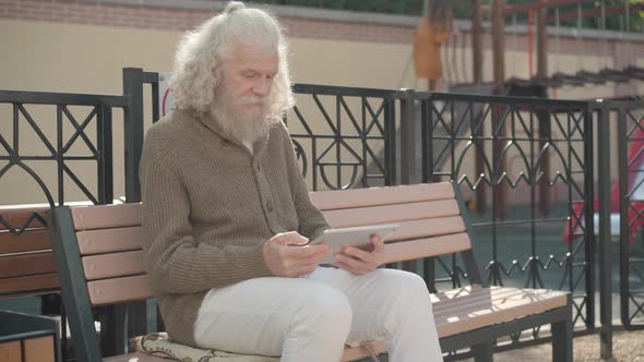 Portrait of Relaxed Modern Senior Man Using Wireless Chat Outdoors with Playground at the Background