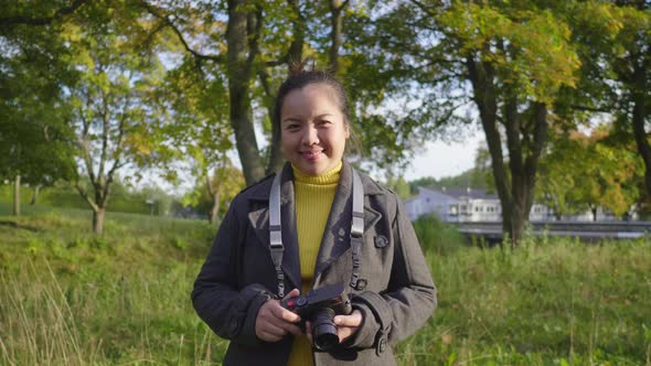 Front view of happy Asian woman standing, holding camera and smiling at forest